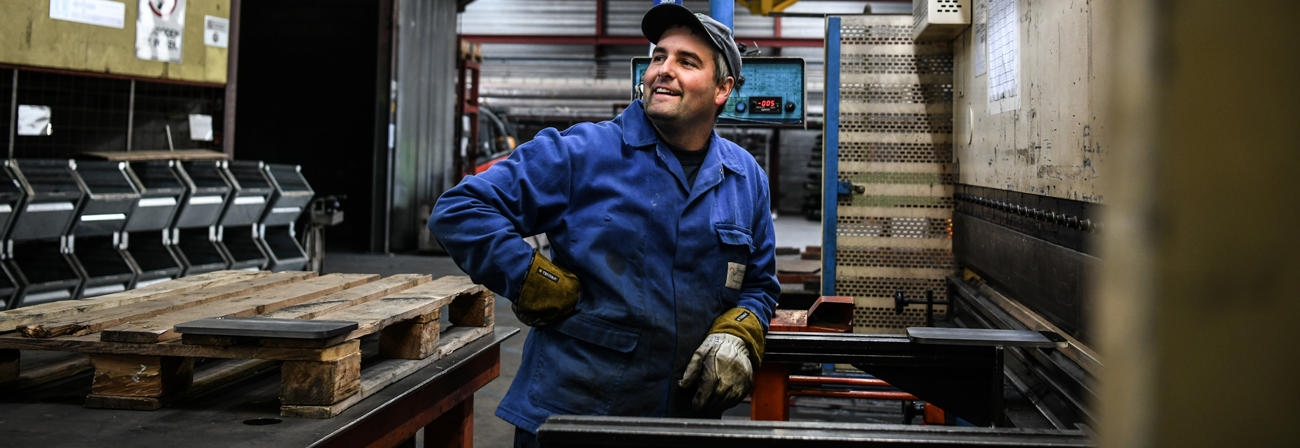 Een allround plaatbewerker is bezig met het bedienen van een plaatschaar in de werkplaats, omringd door diverse gereedschappen en metalen platen. In de achtergrond zijn collega's bezig met andere bewerkingen, zoals slijpen en lassen. De werkplaats is georganiseerd en goed verlicht, wat een professionele en efficiënte werkomgeving weerspiegelt.